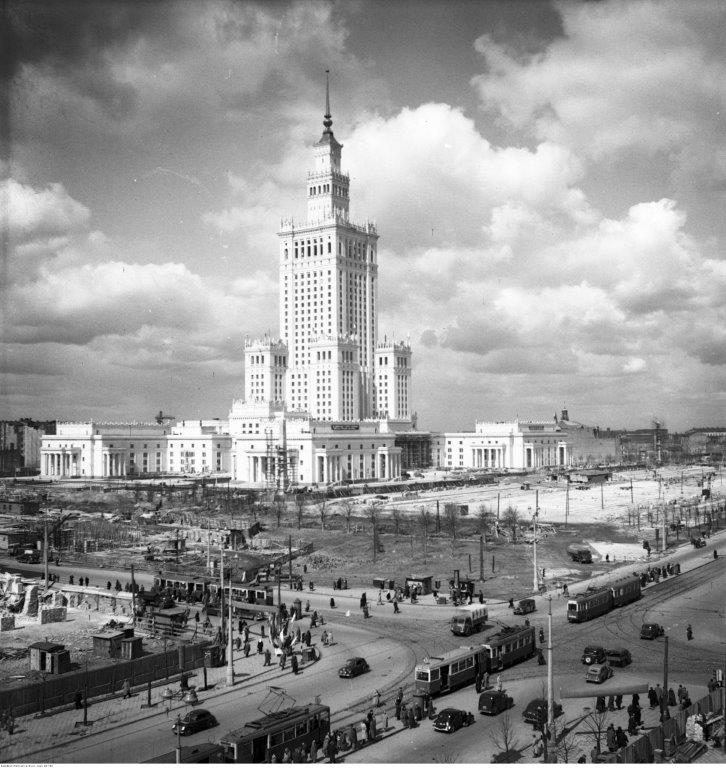 Pałac Kultury wokół pusty plac, na pierwszym planie skrzyżowanie widoczne nieliczne samochody,  tramwaje oraz spore grupy pieszych